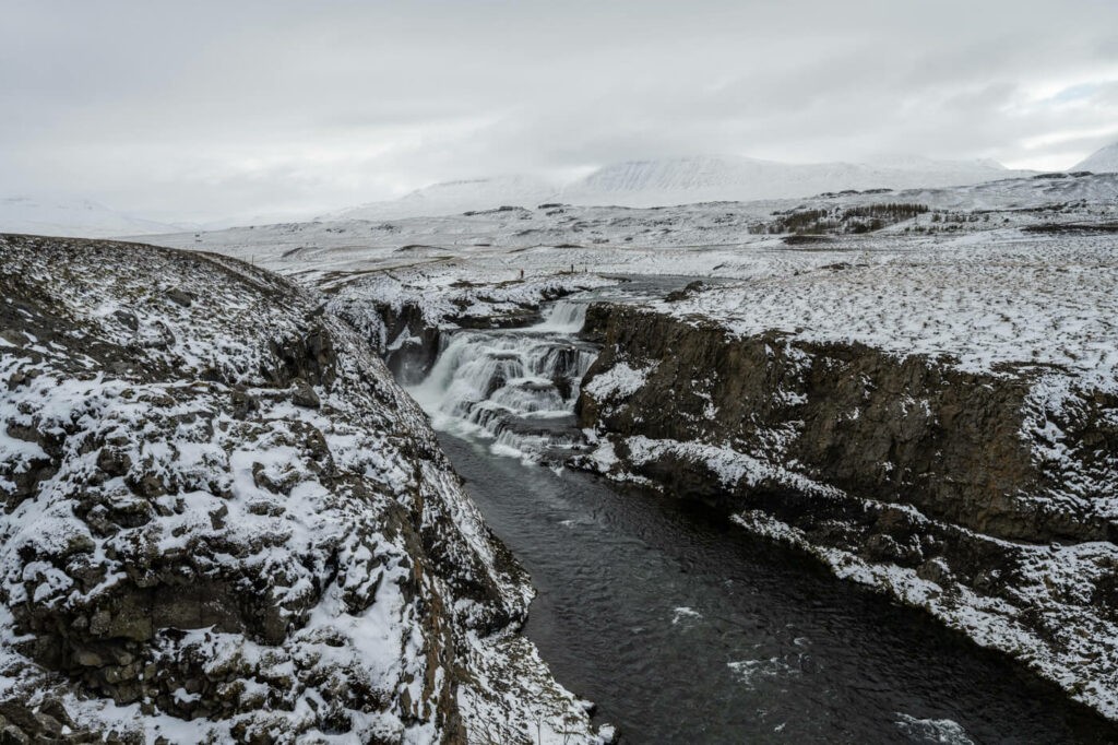 Reykjafoss waterfall viewed from the trail to the Fosslaug Hot Spring on a snow and cloudy day