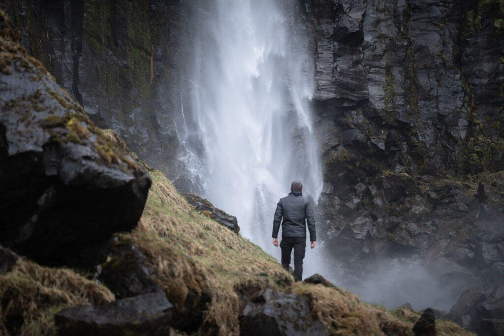 A person dressed in black standing below a waterfall in Iceland. 