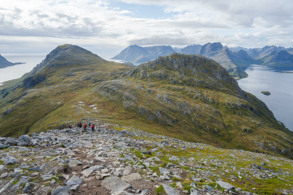 Rocky trail with panoramic views of mountains in a Norwegian fjord and hikers going downhill.
